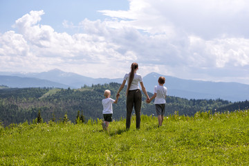 Fototapeta na wymiar Mother and two little sons stand holding hands on a green field against a background of forest, mountains and sky with clouds.