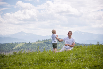 Fototapeta na wymiar Father and young son are blowing dandelions sitting in the grass on a background of green forest, mountains and sky with clouds. Friendship concept