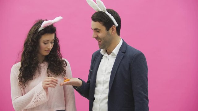 Young couple are beautiful on pink background. During this time, they are dressed in rabble ears. Looking at each other. A man shows carrots and his wife starts to laugh. A young man begins to choke