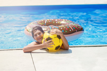 Happy cute little boy teenager lying on  inflatable donut ring with orange in swimming pool.  Active games on water, vacation, holidays concept. Chocolate donut. Cool fun summer holidays for children.