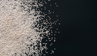 Scattered white sesame seeds on black textured paper background. Close up, copy space, top view....