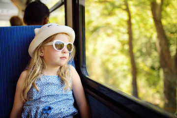 Cute young girl traveling by train on summer day. Child sitting by the window of railway wagon and looking outside.