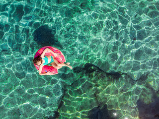 Aerial top down view of cute young girl floating on toy ring at Emplisi Beach, picturesque stony beach in a secluded bay, with clear waters popular for snorkelling.