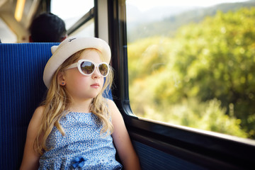 Cute young girl traveling by train on summer day. Child sitting by the window of railway wagon and looking outside.