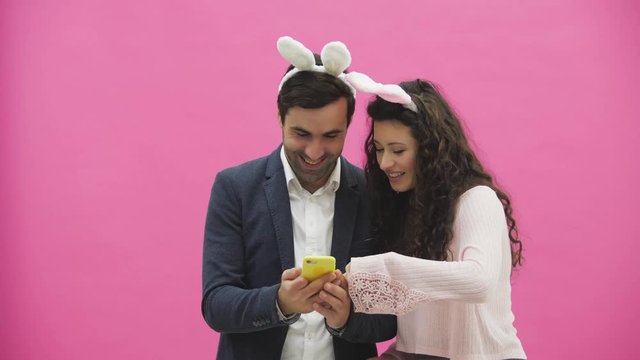 Young lovers couple on pink background. With ravenous ears on the head. During this Easter photo, sephi do on the phone and looking at them rejoice.