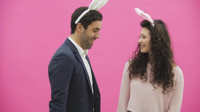 Young sexy couple on pink background. With hackneyed ears on the head. During this man gives a soft toy hare and colored decorative eggs to his wife. Having kissed looking at the camera.