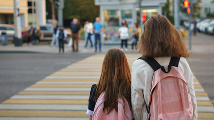 Two cute teen sisters with school backpacks on city street. Girls are standing near pedestrian crossing through highway. Traffic Laws. People are unrecognizable.