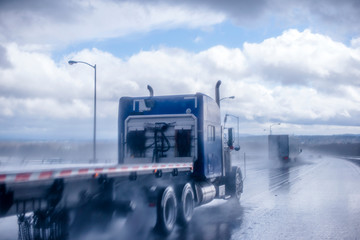Big rig blue classic semi truck with flat bed semi trailer moving on the wet raining road behind another semi truck with rain dust fog