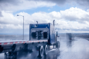 Big rig blue classic semi truck with flat bed semi trailer moving on the wet raining road behind...