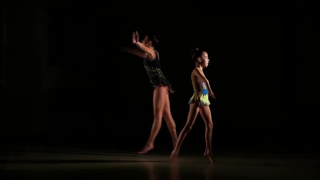 Slow motion, slim cute attractive two girls athletes in bright colorful swimsuits perform elements of rhythmic gymnastics in dark gym with beautiful staged light