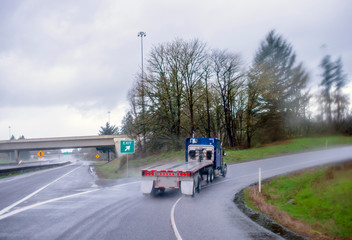 Classic blue big rig semi truck with empty flat bed semi trailer running on the highway exit going uphill in raining weather with wet road