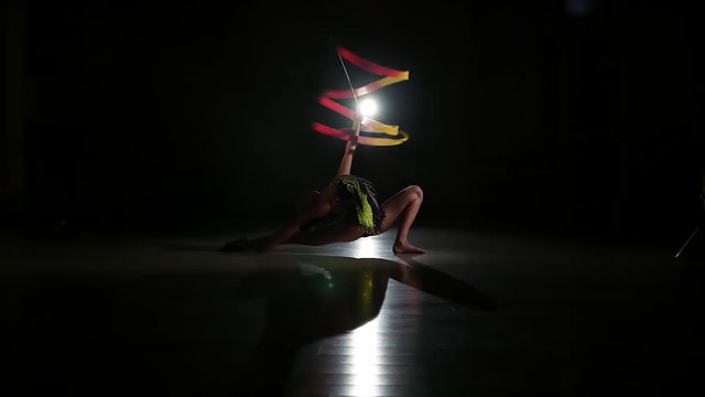 Slow motion, slim cute attractive girl athlete in bright colorful swimsuit performs elements of rhythmic gymnastics with ribbon in dark gym with beautiful staged light and contour light