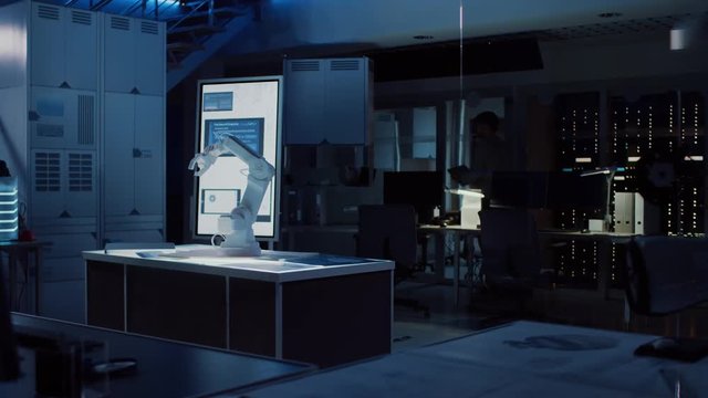 In Industrial Robot Desing Research Laboratory: Robotic Arm Prototype Standing Illuminated on the Desk. In the Dark Background Various High Tech Devices, Computers, Digital Whiteboard and Blueprints