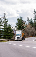 Fototapeta na wymiar White big rig semi truck transporting lumber wood on flat bed semi trailer driving on winding mountain road