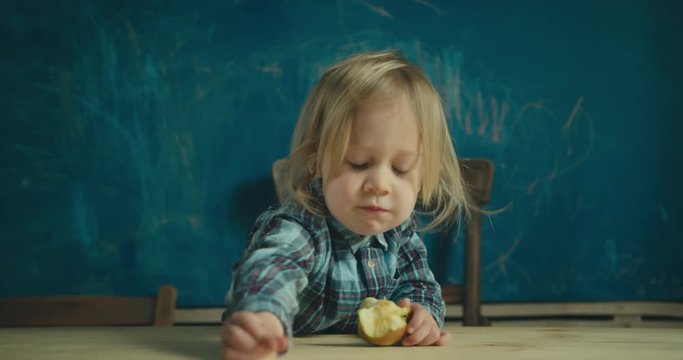 Little toddler eating apple in front of blackboard with his drawings