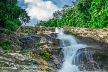 Beautiful waterfall at the mountain with blue sky and white cumulus clouds. Waterfall in tropical green tree forest. Waterfall is flowing in jungle. Nature abstract background. Granite rock mountain.