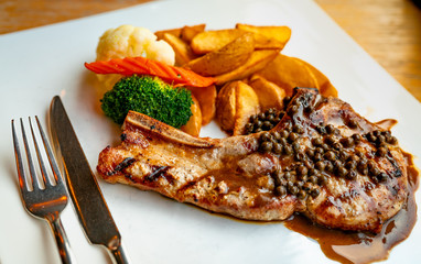 Steak topped with sauce and pepper serve with potato fried and carrots, cauliflower and broccoli steamed. Steak on white plate with stainless steel fork and knife put on wooden table. Tomahawk steak.