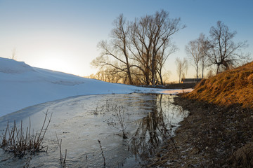A small frozen pond in a ravine in the spring evening. Snow is melting.