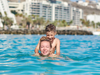 Cute European boy is sitting on his mother’s back, while swimming in the sea.