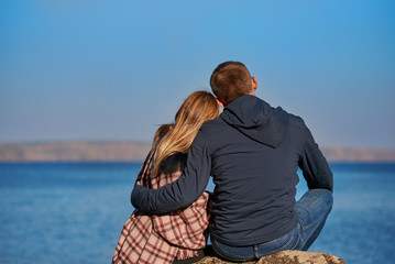 Young beautiful European couple is sitting on the big stone against picturesque lake, they are enjoying the atmosphere around.