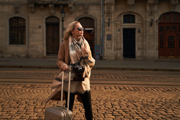 Young beautiful blond woman tourist with a wheeled travel bag and vintage film camera standing on Rynok square in Lviv, Ukraine in the morning