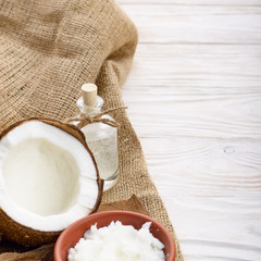 Coconut oil in brown clay bowl, glass bottle, coconut shell with meat on hemp sackcloth on white wooden kitchen table