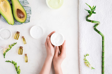 Top view and flat lay of woman holding cream on hands over white table with cosmetic products - avocado oil, cream and bamboo