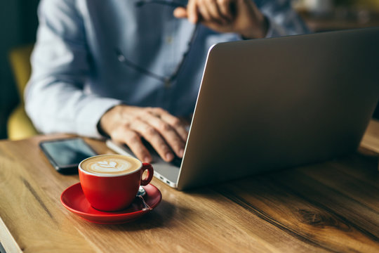 Closeup Of Coffee In Red Mug In Cafeteria. Blurred Man Using Laptop In Background