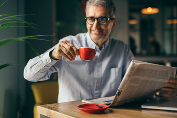 mature gray haired man drinking coffee and reading newspaper in cafe bar