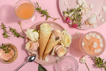 Light pink set of dishes on the table. Delicate rosebuds of pastel colors and petals on a plate and waffle cones. Top view.