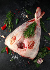 Raw lamb leg on black stone table with herbs. garlic, rosemary, chilli, salt, pepper, Bay leaf.