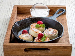 Breakfast in a pan on a white background. Cheesecakes with raspberries and sour cream. On a wooden tray.