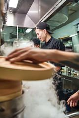 Obraz na płótnie Canvas Cooking process. Close up of chef's hand holding wooden lid while preparing food with his two assistants in a restaurant kitchen