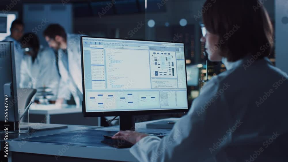 Fototapete Over the Shoulder Shot: Female IT Scientist Uses Computer Showing System Monitoring and Controlling Program. In the Background Technology Development Laboratory with Scientists, Engineers Working 