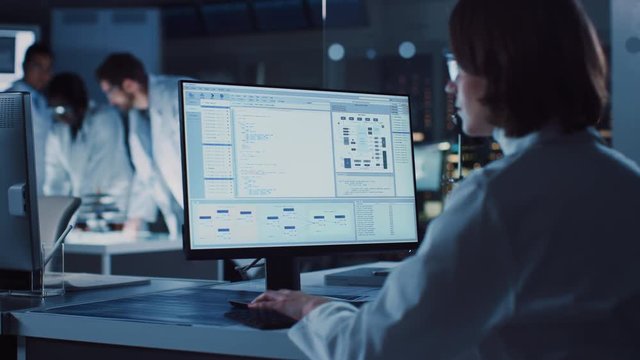 Over the Shoulder Shot: Female IT Scientist Uses Computer Showing System Monitoring and Controlling Program. In the Background Technology Development Laboratory with Scientists, Engineers Working 