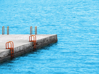 Ladder to the water on the beach. Rusty orange stairs for descending into the blue sea on a concrete pier. Grab bars ladder with blue water in the background