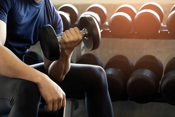 Handsome man playing biceps with dumbbell exercise to strong arm and body sit on black bench at gym fitness background.