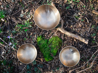 Three singing bowls sitting on soil outdoors around a green heartshaped moss stone.