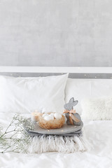 White modern bedroom with Easter decoration. Bed with white bedding set, concrete tray, nest with white eggs, decorative bunny figure, candle and  gypsophila flowers.