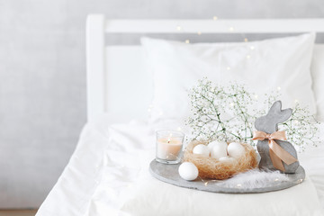 White modern bedroom with Easter decoration. Bed with white bedding set, pillows, concrete tray, nest with white eggs, decorative bunny figure, candle and  gypsophila flowers.
