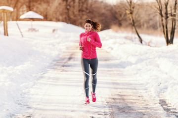 Smiling beautiful Caucasian woman in sportswear running on snowy weather and listening to the music. Wintertime, healthy lifestyle concept.