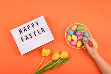 Easter composition, greeting card with child hand, lightbox text Happy Easter, yellow tulips, colored decorative eggs on color background.