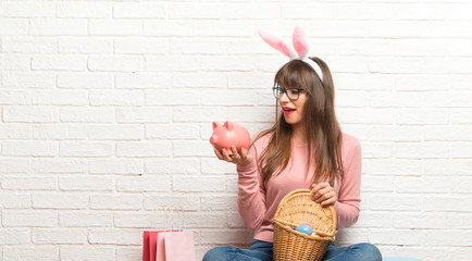 Woman with bunny ears for Easter holidays sitting on the floor surprised while holding a big piggybank