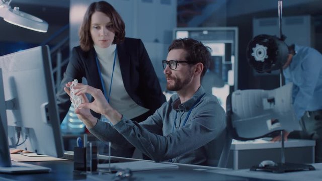 Machinery Engine Development Engineer Working on Computer at His Desk, Talks with Project Colleague, Shows Her Prototype Component. Team of Professionals Working in Modern Industrial Design Laboratory