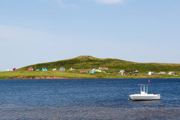 Fototapeta na wymiar Small white boat anchored in water with scattering of colorful houses on Magdalen Islands shore, Quebec, Canada 