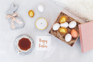Easter decoration. Nest with white and gold eggs in pink box, decorative bunny figure made of concrete, candle, cup of tea, white fluffy plaid and card with greeting Happy Easter on grey background.