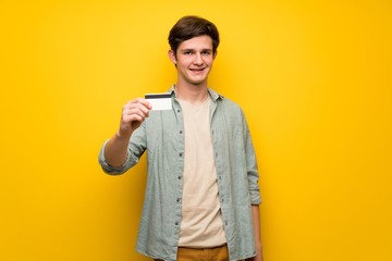 Teenager man over yellow wall holding a credit card
