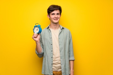 Teenager man over yellow wall holding vintage alarm clock