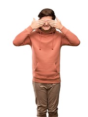 Teenager man with sweatshirt covering eyes by hands. Surprised to see what is ahead over isolated white background