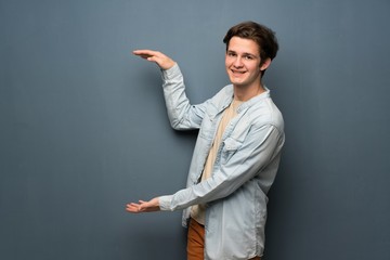 Teenager man with jean jacket over grey wall holding copyspace to insert an ad